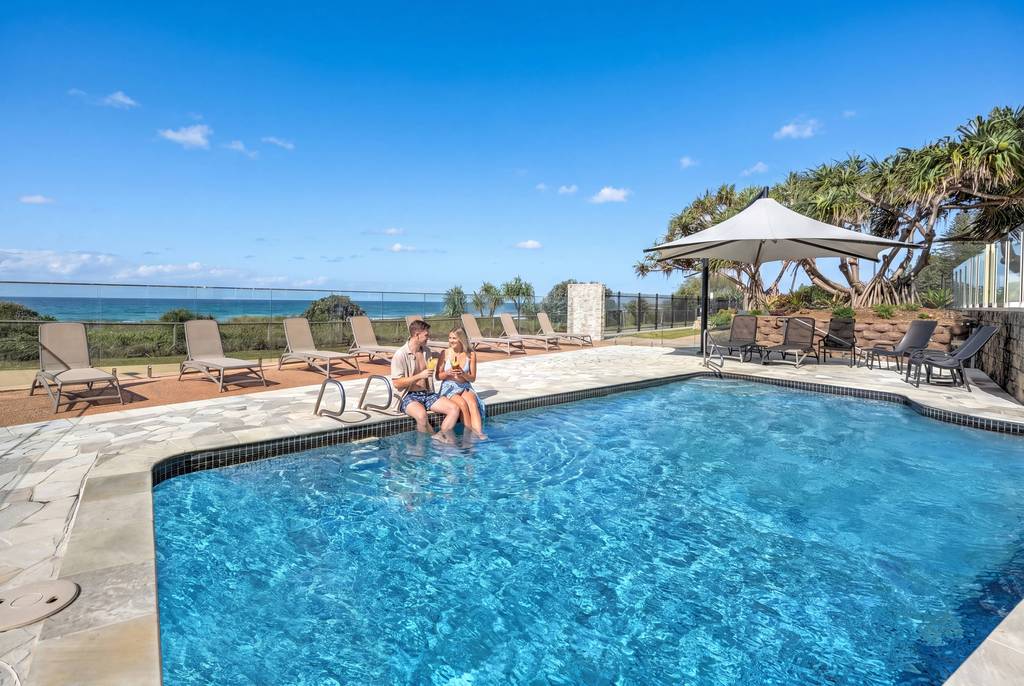 Outdoor heated pool with lounge chairs, umbrellas and ocean beyond the palms